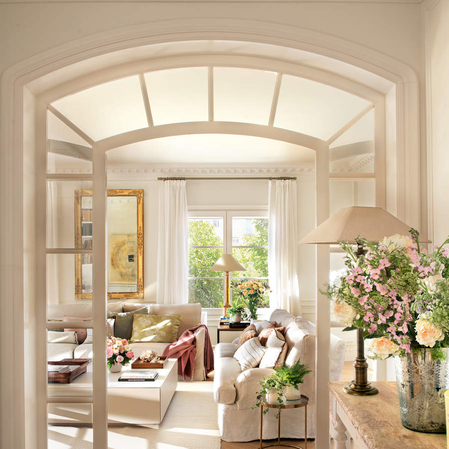 Recibidor clásico con salón al fondo, puerta con arco de cristal, sofás blancos y flores